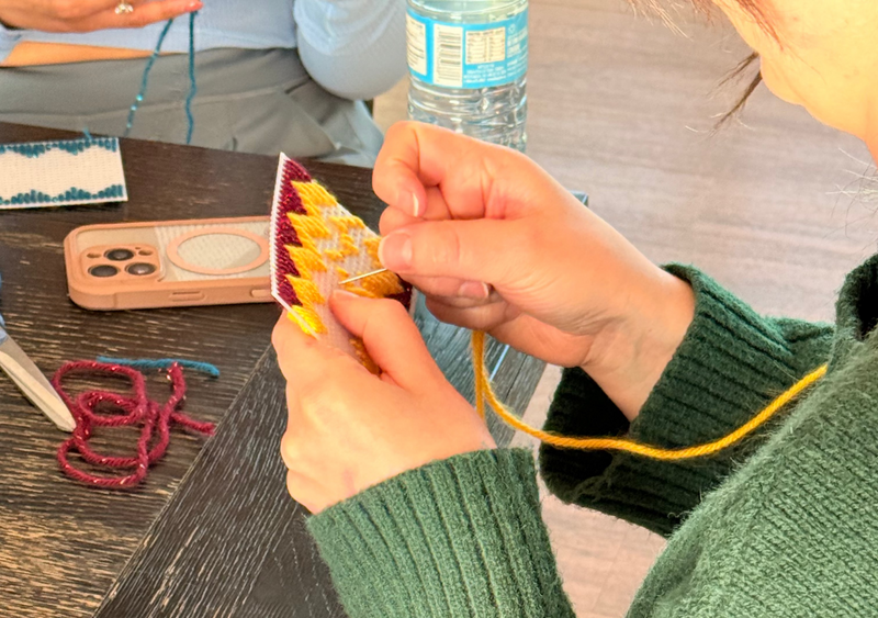 A close-up lifestyle shot of a person wearing a green cable-knit sweater, holding a small yellow and red patterned card or envelope while threading a yellow string. They are seated at a dark wooden table with a water bottle and other items in the background.